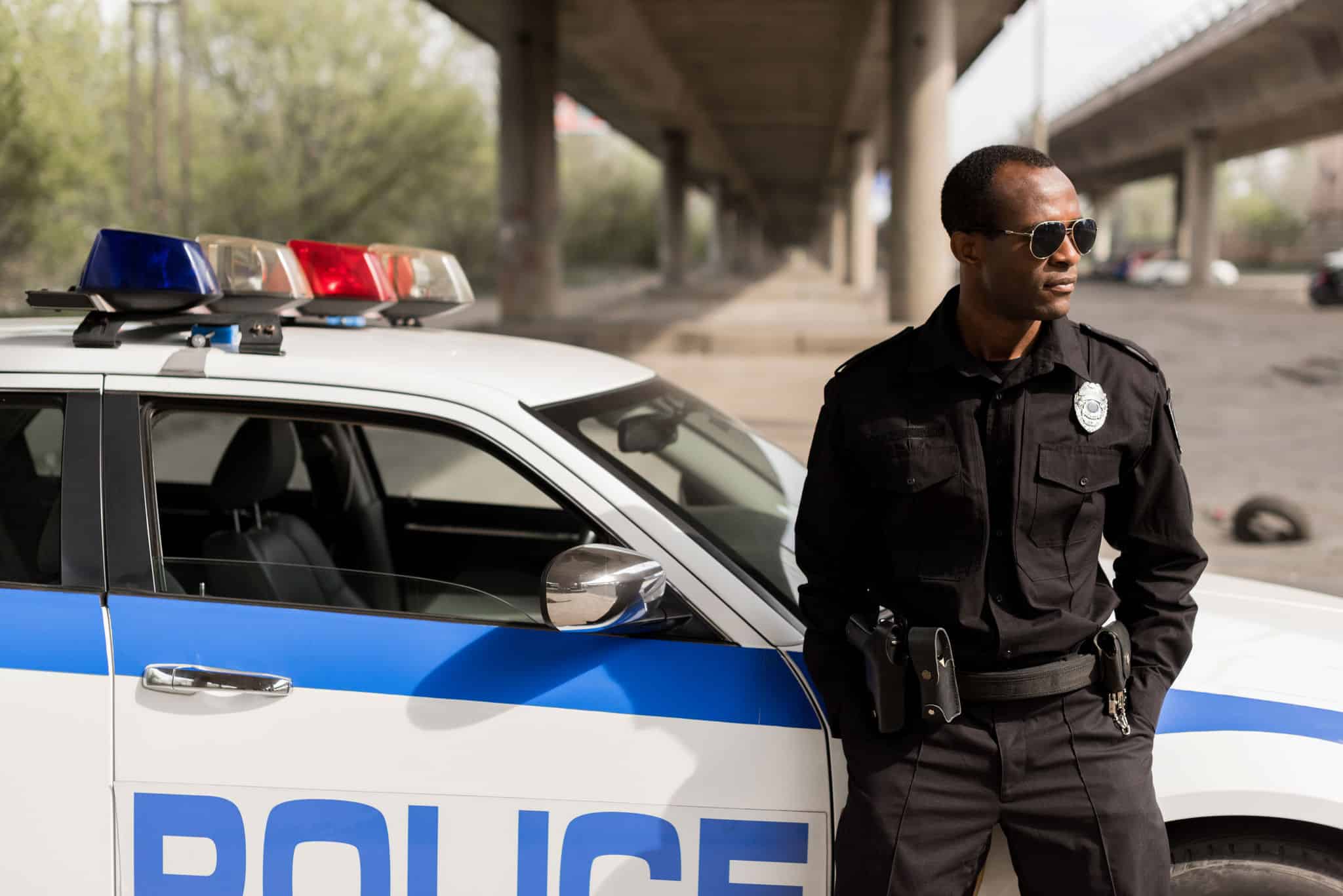 african american police officer leaning back on car and looking away