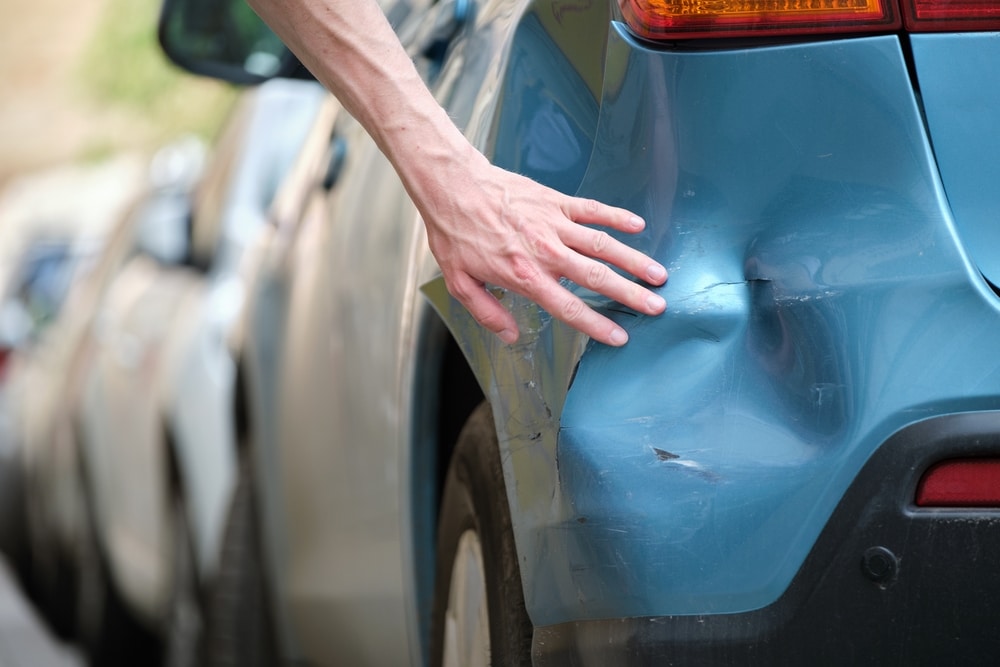 Hand reaching toward a large dent on the rear fender of a blue sedan after it was hit on the street